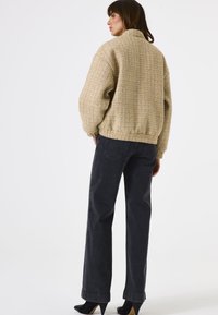 Femme portant une veste beige texturée, un jean noir à jambes larges et des escarpins noirs à bouts pointus, se tenant de dos sur fond blanc.