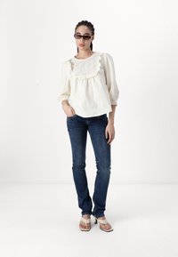 Blusa blanca con cuello bordado y ribetes con volantes, combinada con vaqueros de mezclilla azul oscuro y sandalias de tacón de color claro.