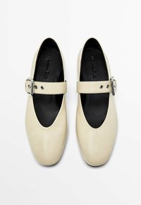 Beige læderballerinaer med rund tå, der har en bred rem med en sølvspænde og sort inderside. Glat tekstur.