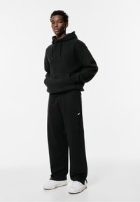 Sort hættetrøje med kængurulomme, snoretræk i hætten, kombineret med sorte sweatpants med et lille logo; hvide sneakers fuldender outfittet.