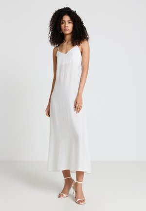 Donna con capelli ricci che indossa un lungo vestito bianco senza maniche e sandali bianchi con tacchi alti, in piedi davanti a uno sfondo semplice.