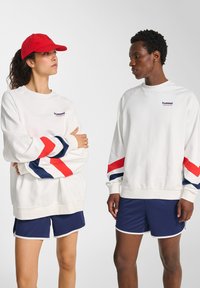 Twee modellen dragen witte sweatshirts met rode en blauwe mouwstrepen en marineblauwe shorts; één model draagt een rode pet en staat tegen een effen achtergrond.