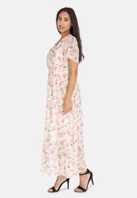 Vestido maxi floral de tela rosa claro con detalles en verde. Presenta mangas cortas, cintura fruncida y un corte holgado. Combinado con tacones negros.