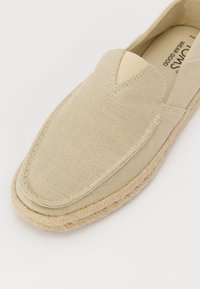 Zapato slip-on de lienzo beige con punta redondeada y detalle de cuerda trenzada a lo largo de la suela. Presenta una costura y un diseño de corte en forma de V en la parte delantera.