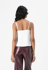 Camicetta in satin bianco con spalline sottili, caratterizzata da un retro dritto e una texture liscia, abbinata a pantaloni larghi bordeaux lucidi.