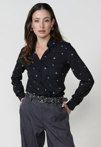 Žena s dugom smešnom kosom pozira samouvereno, noseći crnu majicu s belim uzorcima i sive hlače, uz dodatak remena s uzorkom zmije.