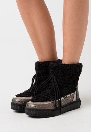 Bottes basses noires texturées avec bordure métallique, détails cloutés, semelles épaisses et lacets noirs portées sur des jambes nues.