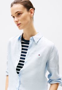 Lyseblå lin skjorte med knepping krage, rullede ermer og liten logo aksent. Brukt over en marineblå og hvit stripet topp.