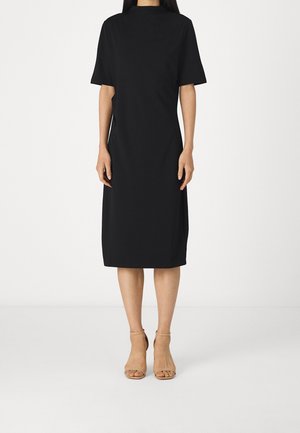 Žena v černých krátkých šatech po kolena a béžových sandálech s otevřenou špičkou na vysokém podpatku, stojící před jednobarevným bílým pozadím.