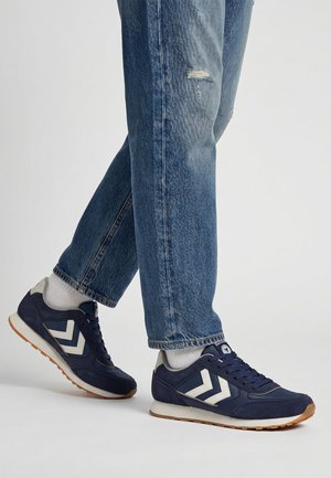 Beine mit leicht abgenutzten blauen Jeans und marineblauen Sneakers mit weißen Chevron-Streifen und weißen Socken stehen auf einem weißen Boden.