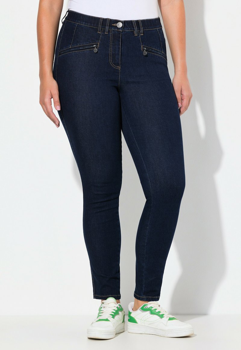 Vaqueros ajustados de color azul oscuro con una cintura alta, dos bolsillos frontales y detalles de metal plateado, combinados con zapatillas blancas con acentos verdes.