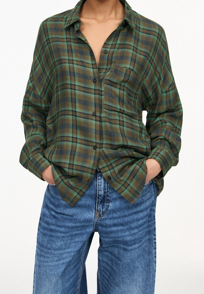 Personne portant une chemise à carreaux verte et marron boutonnée avec manches retroussées et un jean bleu taille haute, mains dans les poches.