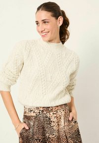 Beiger Strickpullover mit Kabelmuster und hohem Kragen, kombiniert mit einem Rock im Leopardenmuster. Standardpassform mit kurzen, aufgerollten Ärmeln.