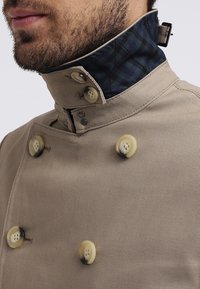Homme portant un manteau beige croisé avec quatre grands boutons et un col relevé doublé d'un tissu à carreaux vert foncé et bleu marine.