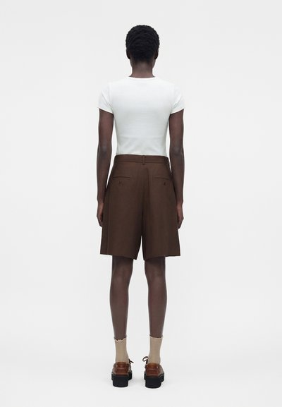 Άτομο που φορά στενό λευκό κοντομάνικο τοπ, καφέ σορτς ως το γόνατο με ψηλή μέση, μπεζ κάλτσες και καφέ παπούτσια με πάτο, στέκεται γυρισμένο με την πλάτη.