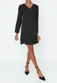 Kvinna iklädd en svart långärmad knälång klänning, svarta spetsiga lägenheter med ankelband och ett silverpärlörhänge, stående på vit golv.