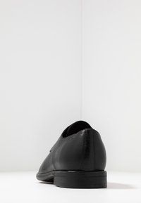 Scarpa in pelle nera con texture liscia, punta arrotondata, tacco basso e design minimalista. Vista posteriore che mette in risalto linee pulite e costruzione.