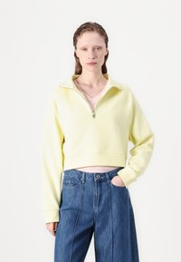 Gekürzter gelber Sweatshirt mit einem Reißverschlusskragen und gerippten Bündchen, kombiniert mit lockeren blauen Jeans aus Denim mit Vorderfalten und Taschen.