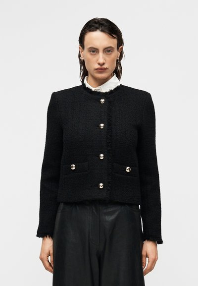 Veste en tweed noir avec un col rond, des boutons à l'avant, deux poches avant et des bords effilochés. Portée sur une chemise blanche à col.