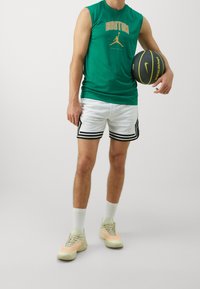 Groene mouwloze shirt met de tekst "BOSTON", witte perforatie-shorts met zwarte bies en lichte sneakers. Houdt een basketbal vast.
