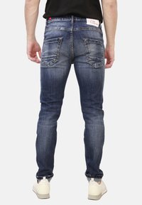 Jeans in denim blu scuro con una vestibilità slim, caratterizzati da tasche posteriori con alette angolate e una lieve scoloritura. Indossati con sneakers bianche.