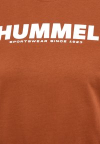 Brun bomull t-shirt med rund hals, med stor vit text som säger "HUMMEL SPORTSWEAR SINCE 1923." Slät textur.