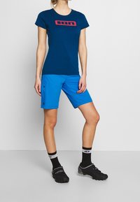 Navy blue short-sleeve t-shirt with red logo, paired with bright blue shorts featuring side pockets. Black cycling shoes and socks complete the outfit.