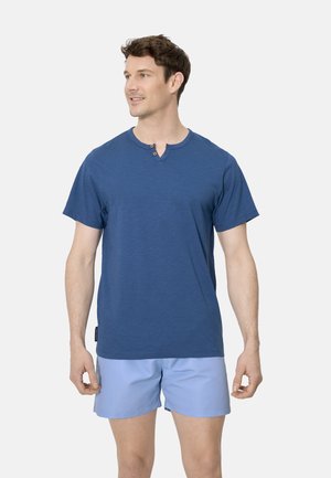 Hombre de pie mirando hacia un lado, vistiendo una camiseta henley de manga corta azul y pantalones cortos azul claro, sobre un fondo blanco.