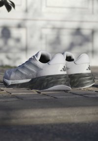 Pari di scarpe da corsa Adidas bianche e nere con il testo "DREAMSTRIKE+", posizionate su una superficie pavimentata all'aperto in una luce naturale soffusa.