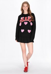 Pulover negru cu text roșu "MERRY" și inimioare roz cu paiete. Purtați cu o fustă plisată neagră și pantofi roșii.
