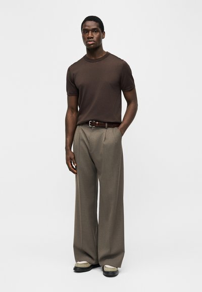 Άνδρας που φοράει καφέ κοντομάνικο πουκάμισο, φαρδιά τζιν παντελόνια ταούπ με πλεκτή ζώνη και μπεζ-μαύρα παπούτσια, στέκεται με το ένα χέρι στην τσέπη.