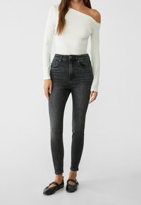 Svarta hög midja skinny jeans i kombination med en vit långärmad topp utan axlar och svarta platta skor, mot en ljus bakgrund.