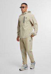 Beige voděodolná bunda s kapucí a zipovou kapsou, v kombinaci s úzkými kalhotami s geometrickým vzorem a logovými akcenty. Stříbrné tenisky.