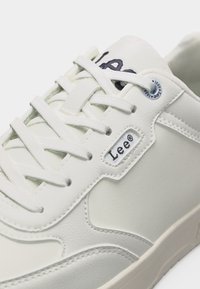 Λευκό δερμάτινο αθλητικό παπούτσι με λευκά κορδόνια, μαύρο έμβλημα "Lee®" στο πλάι και μεταλλικό οπή κορδονιού κοντά στην κορυφή.