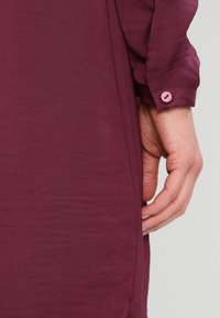 Långärmad skjorta i djup burgunderröd färg med ett strukturerat tyg, som har en rosa knapp på ärmslutet och en sidoklyvning för ökad rörlighet.