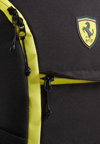 Czarny plecak z żółtymi akcentami, o fakturowanej tkaninie i zamknięciach na zamek. Posiada emblemat Ferrari na przednim panelu. Trwała konstrukcja.