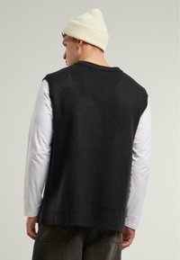 Dark gray sleeveless sweater with a ribbed neckline and hem, paired with a long-sleeve white shirt. Features a casual, relaxed fit.