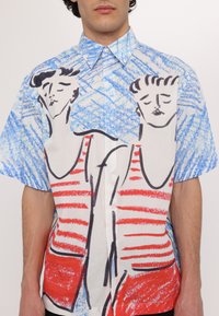 Chemise à manches courtes avec un fond à motif bleu et blanc, ornée de figures rayées rouges entourées de noir. Tissu en coton, coupe décontractée.