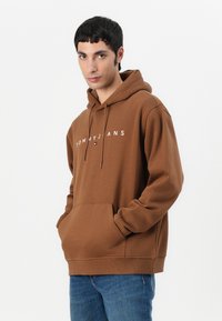 Bruine hoodie met een voorzijde kangoeroezak, trekkoordcapuchon en witte logo-tekst. Gemaakt van zachte, textuurstof met een relaxte pasvorm.