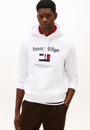 Sweat à capuche blanc avec un logo rouge, bleu et noir et une poche avant, associé à un pantalon marron foncé. Tissu lisse et poignets côtelés.