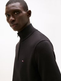 Črna zip-up jopica z visokim ovratnikom, narejena iz pletene tkanine. Ima majhen rdeč, bel in moder logotip na prsih.