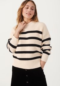 Beige randig tröja med svarta ränder, ribbad textur och hög krage. Bärs med en svart kordflorskjol med knappar fram.