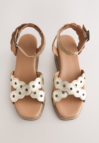 Paire de sandales plates beige avec des brides métalliques dorées festonnées et des boucles à la cheville, bout carré sur un fond neutre.