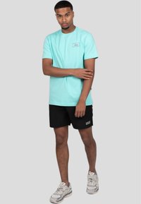 Camiseta de algodón color turquesa claro con cuello redondo y parche de logo. Combinada con pantalones cortos negros y zapatillas deportivas grises. Diseño simple, corte casual.