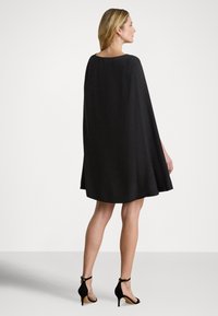 Svart kappekjole med ledig passform, rund halslinning og glatt tekstur. Kombinert med svarte sandaler med hæl og ankelremmer.