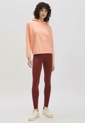 Korallenfarbene Kapuzenjacke mit Kordelzug, kombiniert mit burgunderroten Leggings und cremefarbenen Sneakern. Die Kapuzenjacke hat eine lockere Passform und eine weiche Textur.