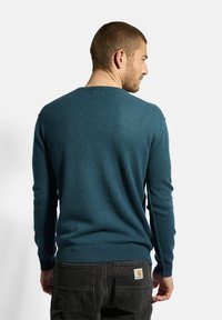 Pullover teal a collo alto realizzato in morbido tessuto testurizzato. Presenta maniche lunghe e polsini a coste, abbinato a pantaloni neri che mostrano un'etichetta.