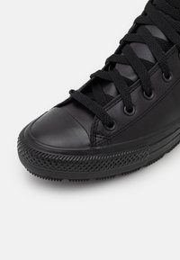 Close-up van een zwarte hoge sneaker met een getextureerde rubberen neus, dikke zwarte veters en een rubberen zool op een witte achtergrond.