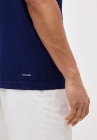 Ανδρικό χέρι δίπλα σε μπλε ναυτικό μπλουζάκι Climacool και λευκό παντελόνι, δείχνοντας υφή υφάσματος και χαλαρή στάση.