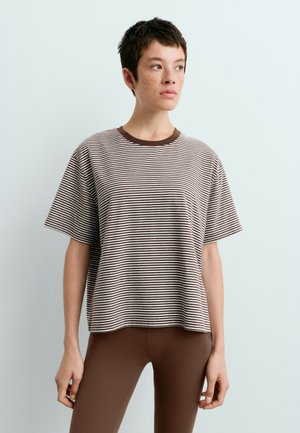 Jong persoon met kort donker haar en sproeten, gekleed in een bruin-wit gestreept T-shirt en effen bruine legging tegen een effen achtergrond.
